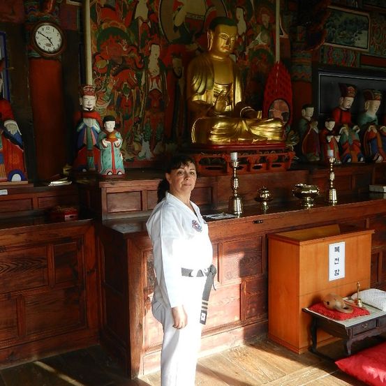 Eine Frau in einem weißen Kampfkunstanzug steht in einem Raum mit einer goldenen Buddha-Statue, Kerzen und verschiedenen Statuen an der Wand. Holzregale und eine Uhr schmücken den Hintergrund.