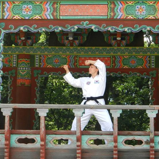 Eine Frau in einem weißen Karate-Anzug mit einem schwarzen Gürtel führt eine Kampfkunstpose auf einem Balkon mit kunstvollen Holzdekorationen durch.