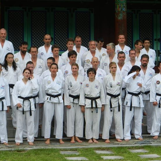 Eine Gruppe von Menschen in weißen Kampfsportuniformen mit schwarzen Gürteln posiert für ein Foto vor einem Gebäude mit grünen Fenstern.