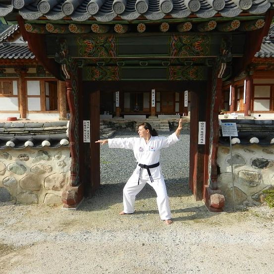 Eine Frau in einem weißen Karate-Anzug steht mit ausgestreckten Armen in einer Kampfkunstpose vor einem traditionellen Gebäude.