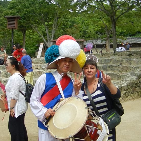 Ein Mann in traditioneller koreanischer Kleidung hält eine Trommel, lächelt und macht ein Friedenszeichen, während eine Frau in einem gestreiften Shirt neben ihm lächelt. Hinter ihnen sitzen Menschen auf Steinstufen, und Bäume und ein Haus sind im Hintergrund zu sehen.