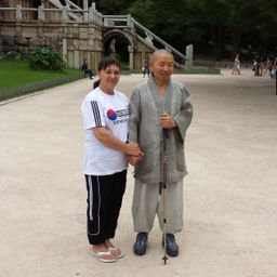 Zwei Personen stehen in einem Park und lächeln in die Kamera. Eine Frau trägt ein weißes T-Shirt mit Tae Kwon Do. Ein Mann in einem Gewand hält einen Stock. Leute gehen hinter ihnen.