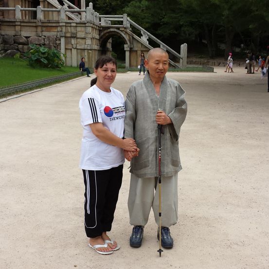 Zwei Personen stehen in einem Park und lächeln in die Kamera. Eine Frau trägt ein weißes T-Shirt mit Tae Kwon Do. Ein Mann in einem Gewand hält einen Stock. Leute gehen hinter ihnen.