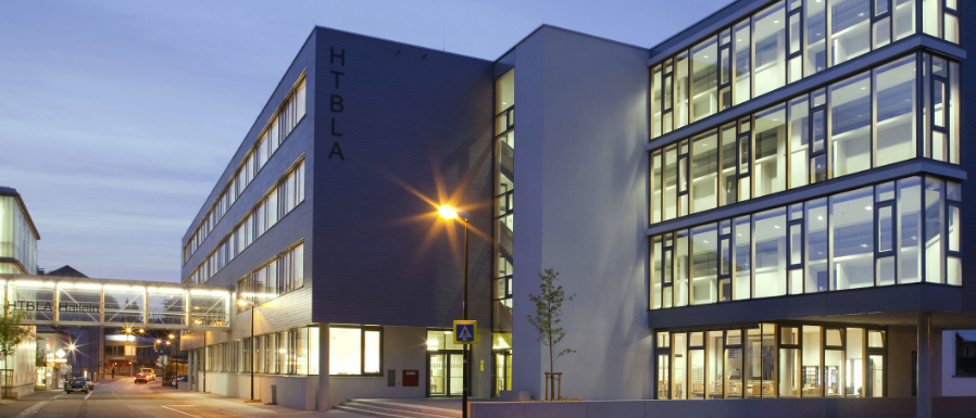 Ein Abendblick auf ein modernes Bürogebäude mit Glasfenstern und einem Baum davor. Das Gebäude ist von Straßenlaternen beleuchtet, und es gibt eine Brücke zum Eingang.