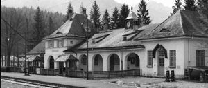 Ein Schwarz-Weiß-Bild eines alten Bahnhofs mit Bogenfenstern und einem Uhrturm auf dem Dach. Im Hintergrund befinden sich Bäume und Berge.
