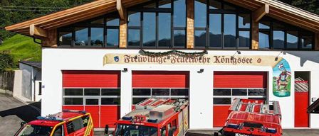 Drei Feuerwehrfahrzeuge vor einem Feuerwehrhaus mit einem Schild, auf dem Freiwillige Feuerwehr Konigssee steht. Im Hintergrund sind Berge zu sehen.