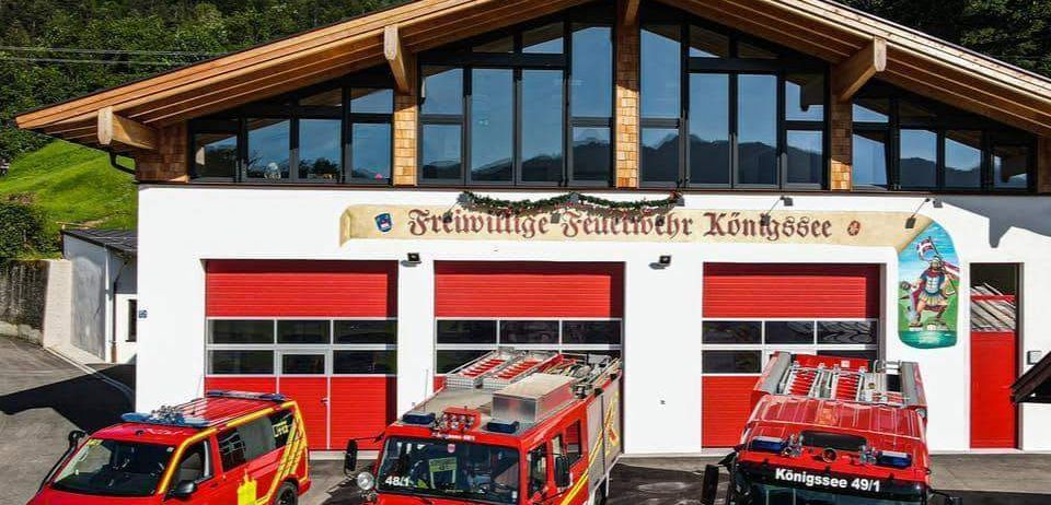 Drei Feuerwehrfahrzeuge vor einem Feuerwehrhaus mit einem Schild, auf dem Freiwillige Feuerwehr Konigssee steht. Im Hintergrund sind Berge zu sehen.