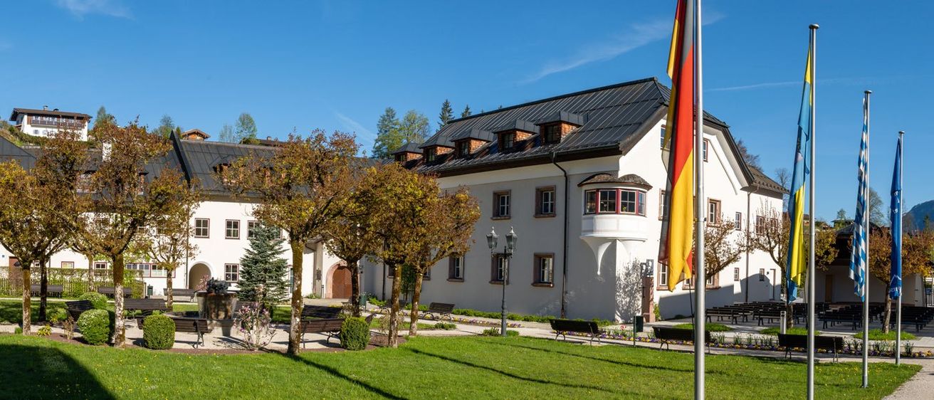 Ein großes weißes Gebäude mit einem grauen Dach und mehreren Fenstern, umgeben von einem Rasen. Bänke und Bäume sind verstreut. Eine deutsche Flagge steht auf einem Pfahl rechts.