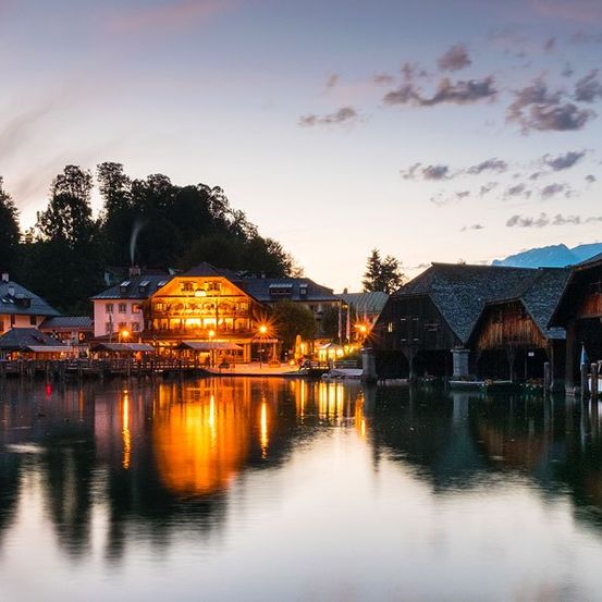Ein Städtchen am See mit mehreren Gebäuden, die am Abend beleuchtet sind. Das Wasser spiegelt die Lichter, und Berge sind im Hintergrund.