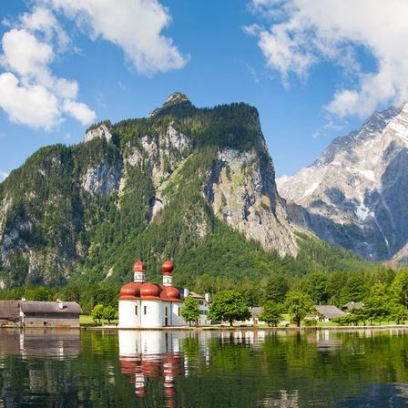 Eine kleine weiße Kirche mit roten Kuppeln steht auf einem See, umgeben von üppigem Grün und Bergen. Das Wasser spiegelt den ruhigen Himmel und die Wolken wider.