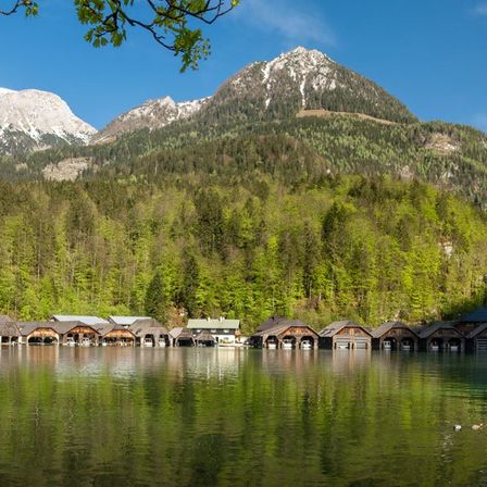Ein ruhiger See mit angedockten Booten und Alpenbergen im Hintergrund unter einem blauen Himmel.