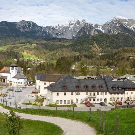 Luftaufnahme eines Dorfes mit einer Kirche und Gebäuden, umgeben von grünen Hügeln und Bergen, mit Autos vor dem Gebäude.
