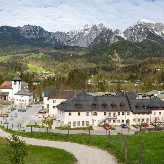 Luftaufnahme eines Dorfes mit einer Kirche und Gebäuden, umgeben von grünen Hügeln und Bergen, mit Autos vor dem Gebäude.