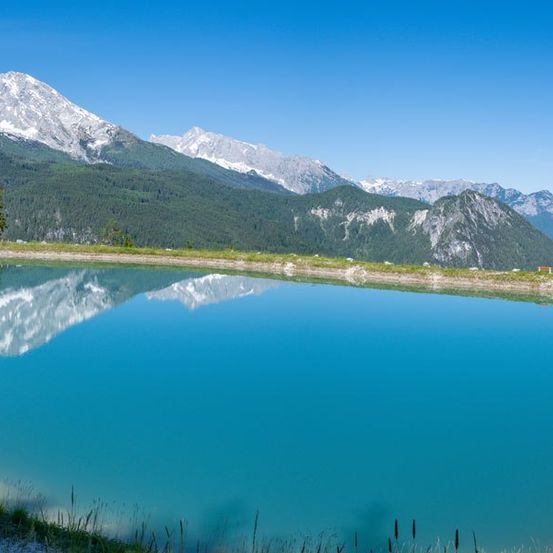 Ein ruhiger Bergsee mit Spiegelungen der schneebedeckten Gipfel unter einem klaren blauen Himmel. Dichte grüne Hügel umgeben das Wasser.