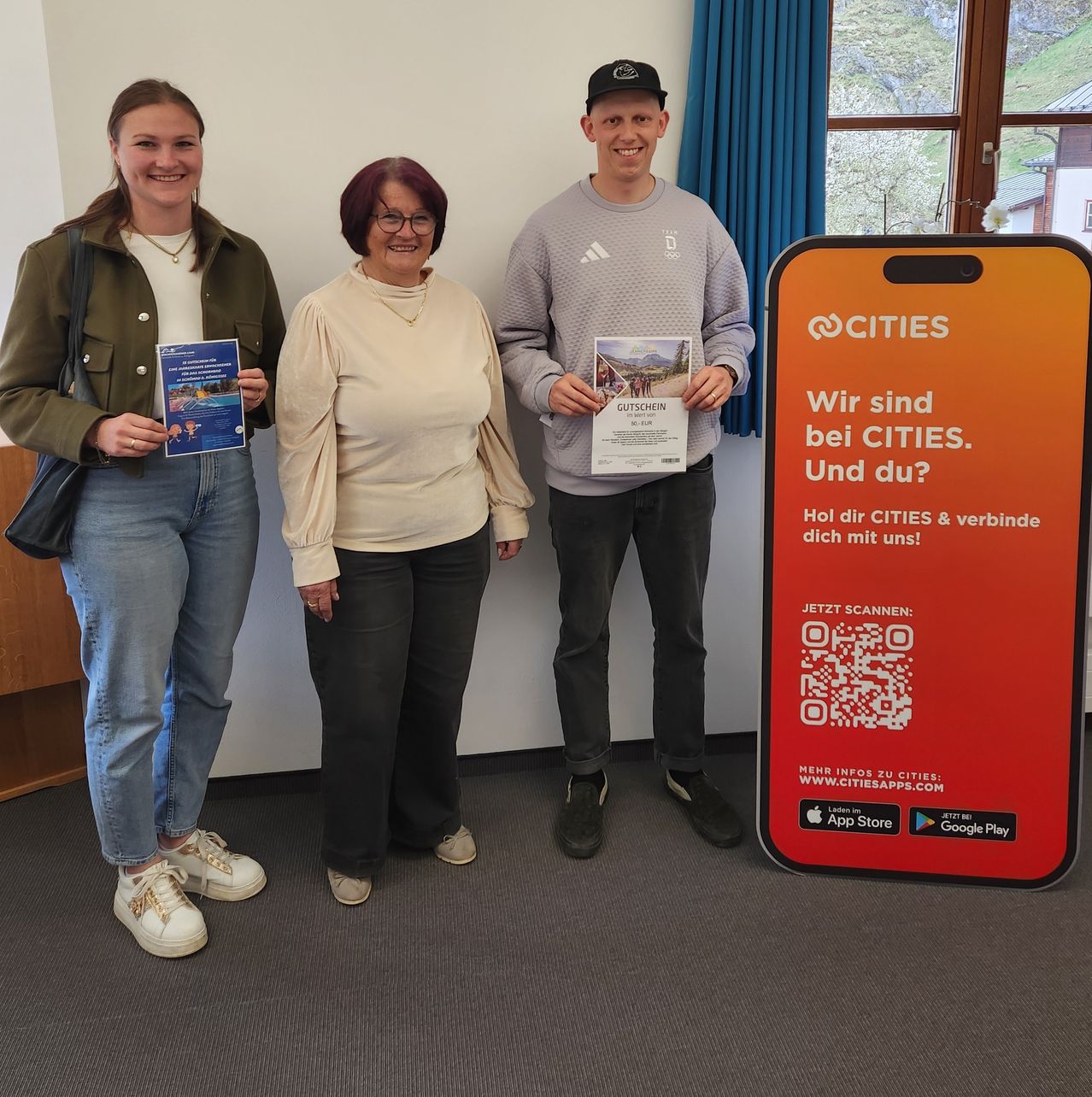 Drei Personen stehen zusammen vor einem Schild, das Cities sagt. Die Frau auf der linken Seite hat ein Buch, die mittlere Frau hat ein Stück Papier und der Mann hat einen Flyer. Sie alle lächeln.