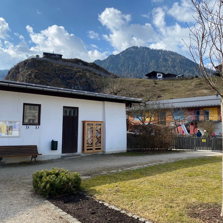 Ein weißes Gebäude mit einer Bank und einem kleinen Baum steht im Vordergrund, mit einem Berg und einem Haus im Hintergrund unter einem blauen Himmel mit Wolken.