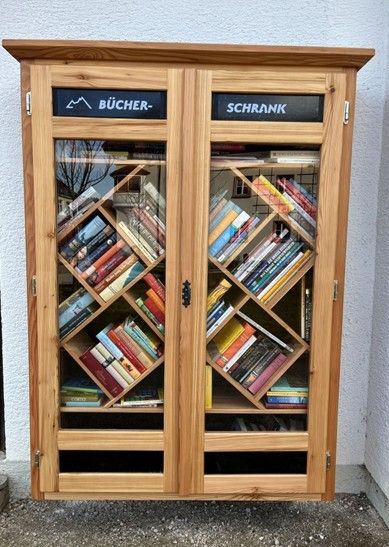 Ein holzernes Bücherregal mit Glastüren. Die linke Tür hat das Schild 'Bücher-' und die rechte 'Schrank'. Es befinden sich viele Bücher darin.