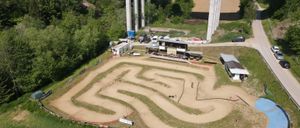 Eine Luftaufnahme einer Schlammrennstrecke mit einem gewundenen Kurs, umgeben von grünem Gras und Bäumen. Fahrzeuge und Strukturen sind in der Nähe, und eine Brücke ist in der Ferne sichtbar.