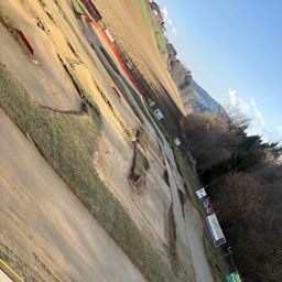 Luftaufnahme einer Motocross-Strecke mit Gras, Bergen in der Ferne und Bannern entlang der Seiten. Die Strecke weist Reifenspuren und eine rote Seilbarriere auf.