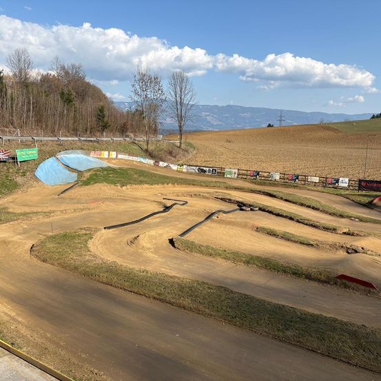 Eine Draufsicht auf eine Schlammpiste mit Kurven, einigen Rampen und Bannern. Sie ist von Gras- und Erdfeldern umgeben. In der Ferne befinden sich Berge und Bäume.