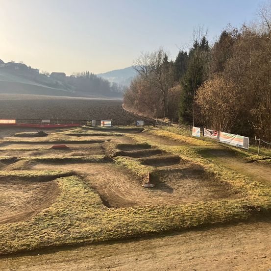 Eine leere Dirtbike-Strecke mit mehreren Kurven. Bäume umgeben die Strecke, und ein Banner ist sichtbar. Die Sonne geht in der Ferne unter.