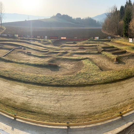 Ein Blick von oben auf eine unfertige Motocross-Strecke mit Kurven und Grasflächen. Die Strecke ist von einer Betonbarriere umgeben. In der Ferne befindet sich ein weitläufiges offenes Feld mit Bäumen und Bergen.