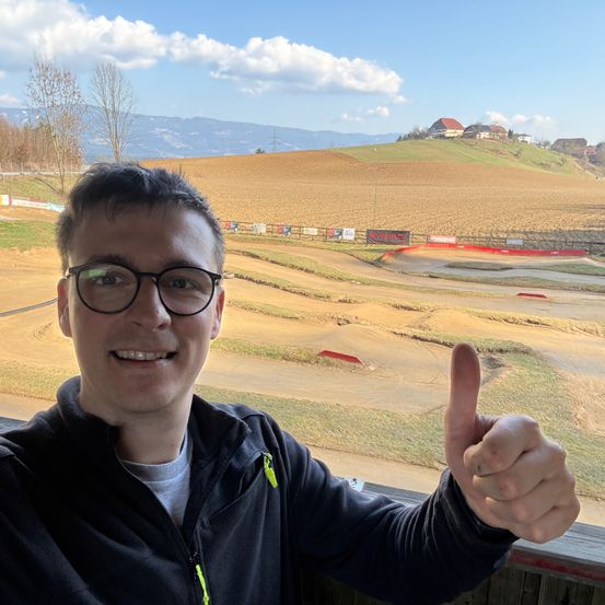 Ein Mann mit Brille lächelt und gibt ein Daumen hoch vor einer Dirt-Rennstrecke. Hinter ihm sind ein grasiges Feld und Berge unter einem teilweise bewölkten Himmel sichtbar.