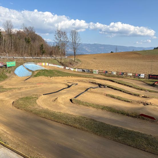Eine Draufsicht auf eine Dirtbike-Strecke mit Kurven und Reifenbarrieren. Im Hintergrund befindet sich ein weites Feld mit Bäumen und Bergen unter einem bewölkten Himmel.