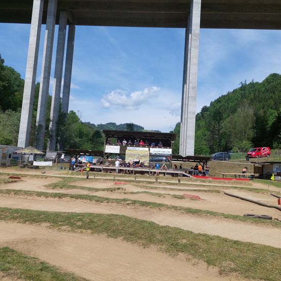 Eine Outdoor-Veranstaltung mit einer Dirt-Piste unter einer Brücke. Zuschauer sitzen in den Tribünen, und Autos sind in der Nähe geparkt. Bäume und ein Berg im Hintergrund.