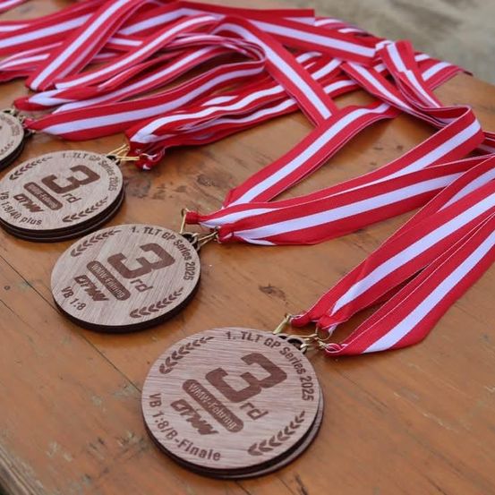 Fünf Holzmedaillen, beschriftet mit 1. bis 3., sind auf einer Holzfläche mit roten und weißen Bändern ausgestellt. Sie sind Teil der TLT GP Serie 2025.