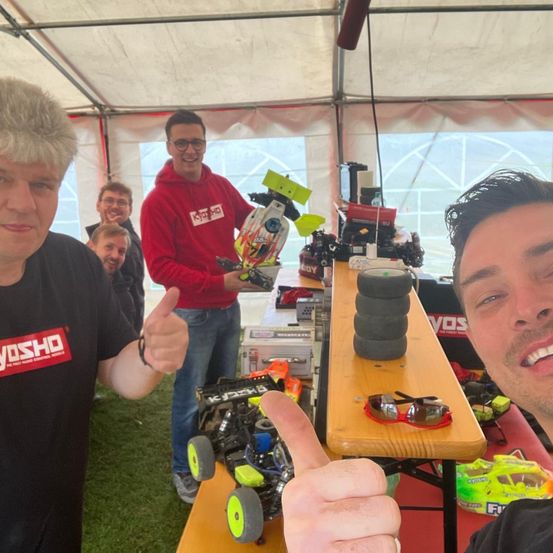 Zwei Männer, die Daumen hoch halten vor einem Tisch voller RC-Autos, einer in einer roten Kapuze und der andere in einem schwarzen Shirt mit Yosho-Logo.