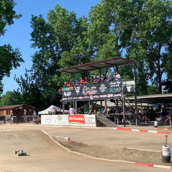 Ein Rennen mit ferngesteuerten Autos auf einer Erdstrecke. Zuschauer sitzen auf den Tribünen. Ein Banner mit verschiedenen Sponsoren und Logos ist sichtbar. Bäume und ein klarer Himmel bilden den Hintergrund.