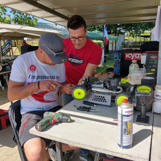 Zwei Männer arbeiten an einem Automodell, einer mit einer Bohrmaschine, der andere mit einem Schraubenzieher. Sie sind unter einem Vordach mit verschiedenen Werkzeugen und Ausrüstungsgegenständen.