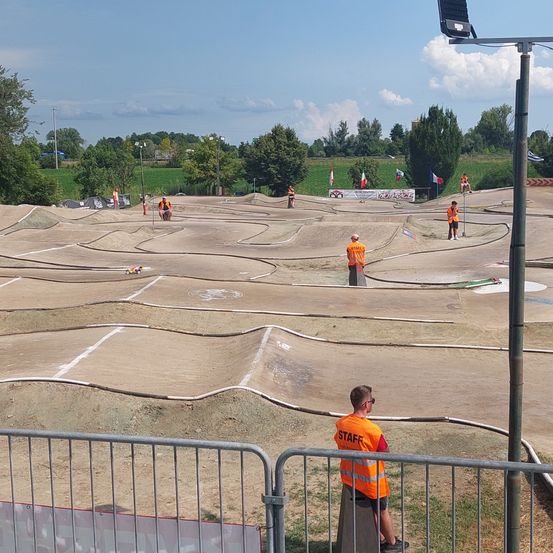 Eine Dirt-Bike-Rennstrecke mit mehreren Fahrern in orangefarbenen Uniformen. Einige gehen, andere stehen. Zuschauer beobachten von hinter Metallzäunen aus.
