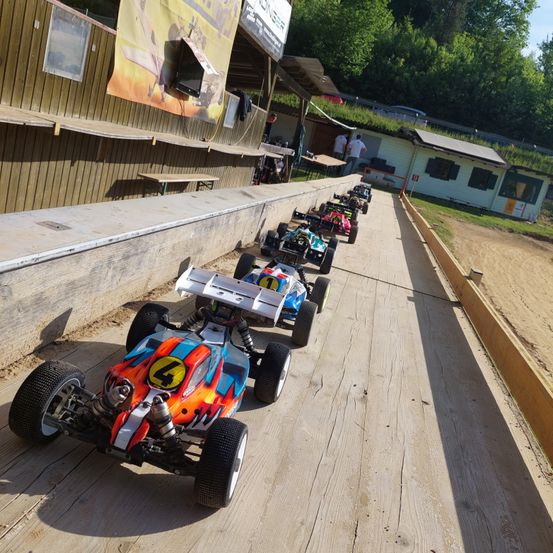 Eine Reihe bunter Spielzeugautos mit Nummern stehen auf einer Holzbahn. Im Hintergrund befindet sich ein Gebäude mit grünem Dach und einem Banner.