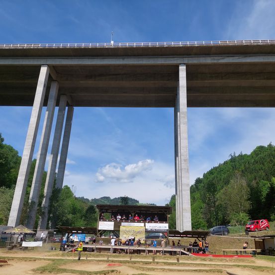 Ein Dirtbike-Wettkampf findet an einem sonnigen Tag unter einer Brücke statt. Zuschauer sehen von einer Plattform zu, während ein rotes Auto und ein Pickup-Truck in der Nähe geparkt sind.