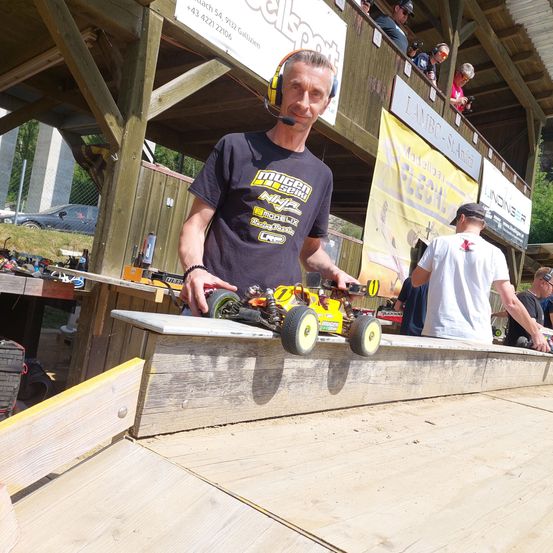 Ein Mann mit Kopfhörern posiert mit einem gelben ferngesteuerten Auto auf einer Rampe bei einer Rennveranstaltung. Andere Rennfahrer und Zuschauer sind im Hintergrund.