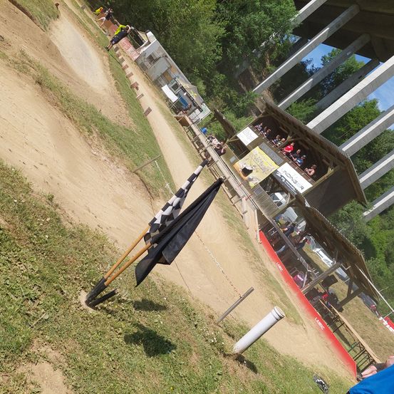 Ein Rennen auf einer Schotterpiste mit einer schwarz-weißen Fahne und einem Rennwagen auf der Strecke. Es gibt Zuschauer, die von einer Struktur aus zusehen. Bäume und eine Brücke sind im Hintergrund.