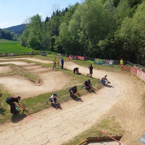 Eine Gruppe von Menschen bereitet sich auf ein Dirtbike-Rennen auf einer Rennstrecke vor. Zwei Personen sind gebückt und stellen die Fahrzeuge ein. Mehrere andere stehen oder knien auf den Rasenflächen.