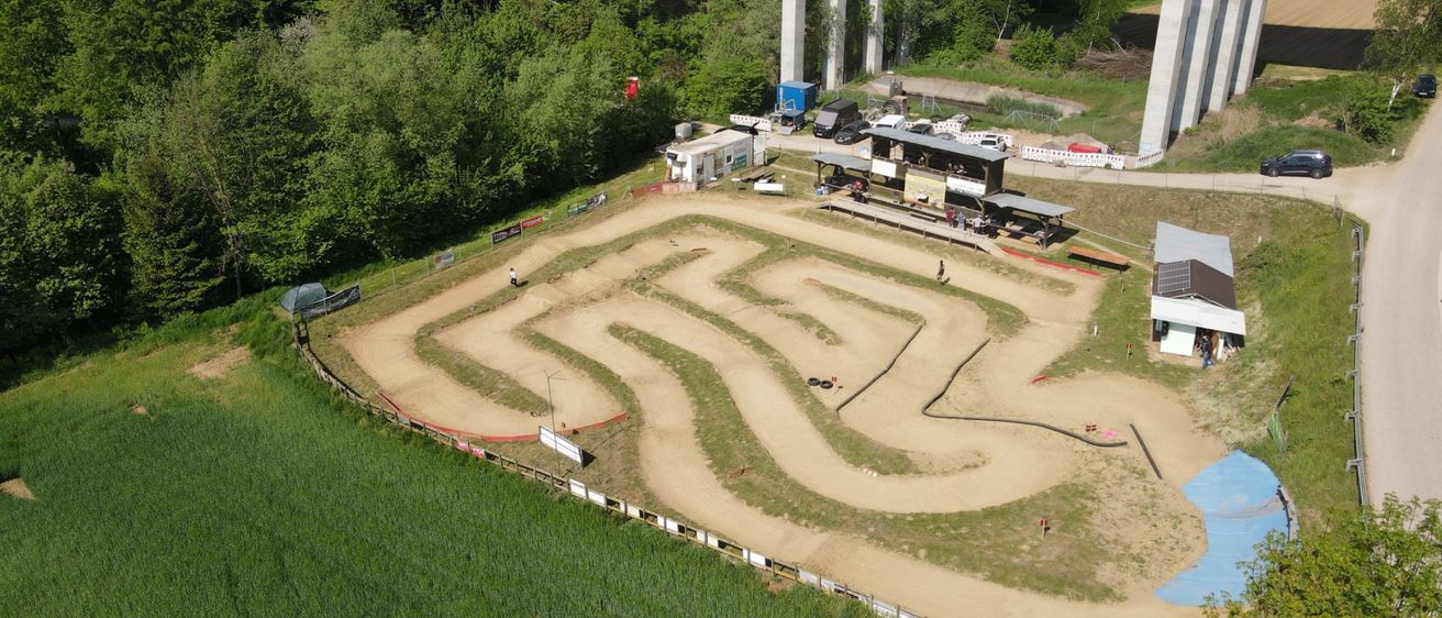 Eine Draufsicht auf eine Motocross-Strecke mit mehreren Fahrzeugen und Personen, vor dem Hintergrund von Bäumen und einer Brücke.