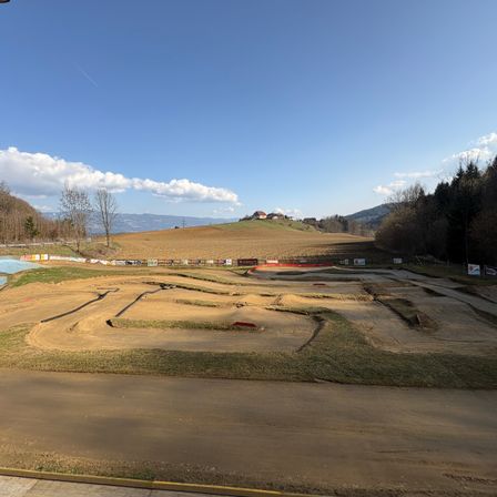 Ein weiter Blick auf eine unfertige Rennstrecke mit Erde, Gras und Zäunen. In der Ferne ist ein Hügel mit Bäumen und einem Gebäude unter einem blauen Himmel mit Wolken sichtbar.