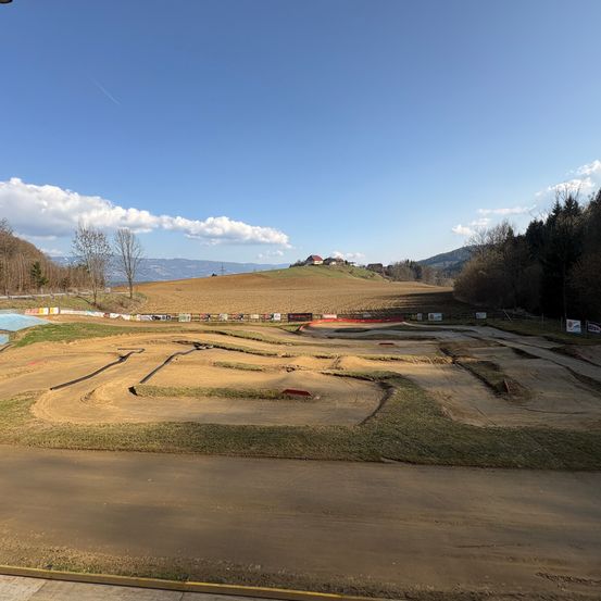 Ein weiter Blick auf eine unfertige Rennstrecke mit Erde, Gras und Zäunen. In der Ferne ist ein Hügel mit Bäumen und einem Gebäude unter einem blauen Himmel mit Wolken sichtbar.