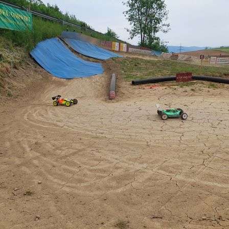 Zwei ferngesteuerte Autos rasen auf einer trockenen Erdbahn. In der Nähe befinden sich blaue Planen, ein Rohr und ein Stoppschild.