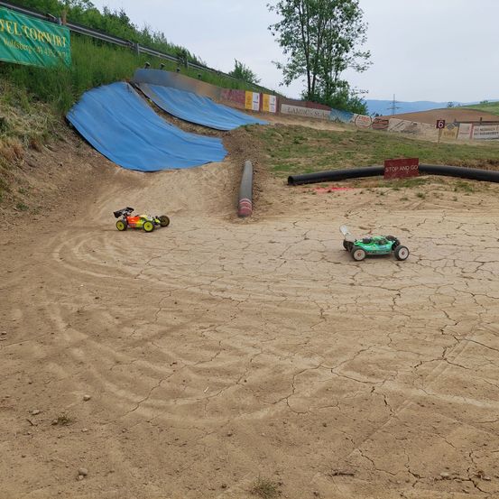 Zwei ferngesteuerte Autos rasen auf einer trockenen Erdbahn. In der Nähe befinden sich blaue Planen, ein Rohr und ein Stoppschild.
