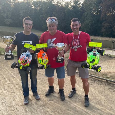 Drei Männer stehen auf einem Erdweg und halten Pokale und ferngesteuerte Autos in den Händen. Der Mann auf der linken Seite trägt eine Brille und ein schwarzes T-Shirt. Der Mann in der Mitte trägt ein rotes T-Shirt und hat eine Medaille. Der Mann auf der rechten Seite trägt ein rotes T-Shirt und Shorts. Bäume und eine Wand sind im Hintergrund.
