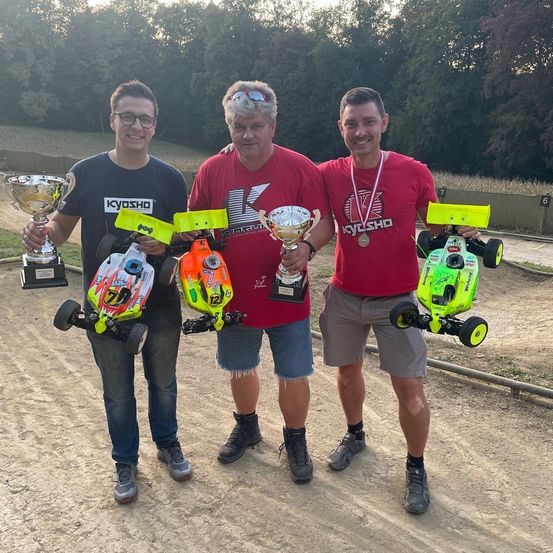 Drei Männer stehen auf einem Erdweg und halten Pokale und ferngesteuerte Autos in den Händen. Der Mann auf der linken Seite trägt eine Brille und ein schwarzes T-Shirt. Der Mann in der Mitte trägt ein rotes T-Shirt und hat eine Medaille. Der Mann auf der rechten Seite trägt ein rotes T-Shirt und Shorts. Bäume und eine Wand sind im Hintergrund.
