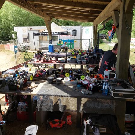 Ein Freiluftaufbau mit Tischen unter einem Vordach, gefüllt mit RC-Autos, Werkzeugen und verschiedenen Gegenständen. Ein Mann arbeitet an einem Tisch, mit einem weißen Zelt und einem Auto im Hintergrund.