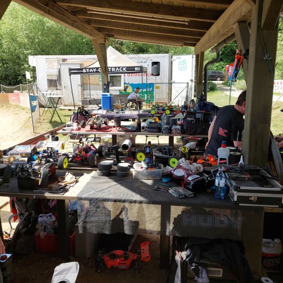 Ein Freiluftaufbau mit Tischen unter einem Vordach, gefüllt mit RC-Autos, Werkzeugen und verschiedenen Gegenständen. Ein Mann arbeitet an einem Tisch, mit einem weißen Zelt und einem Auto im Hintergrund.