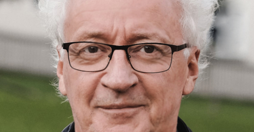 Ein Mann mit weißem Haar, der eine Brille und eine schwarze Jacke trägt, lächelt und schaut nach vorne.