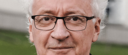 Ein Mann mit weißem Haar, der eine Brille und eine schwarze Jacke trägt, lächelt und schaut nach vorne.